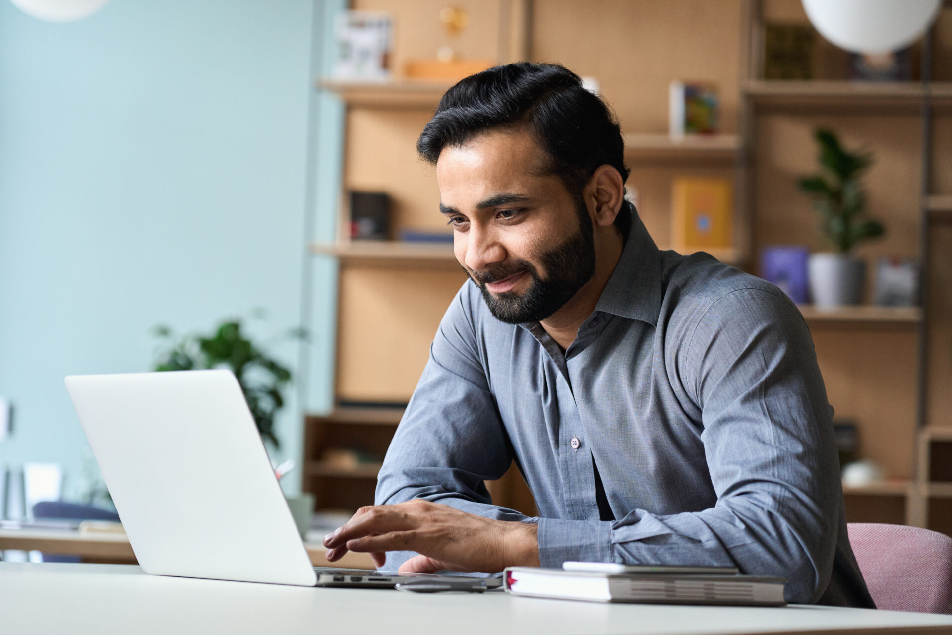 Smiling indian business man working on laptop at home office. Young indian student or remote teacher using computer remote studying, virtual training, watching online education webinar at home office.